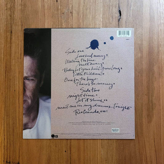The back cover of a vinyl record album, featuring a handwritten tracklist and a partial portrait of a man on the left, displayed flat on a wooden surface.