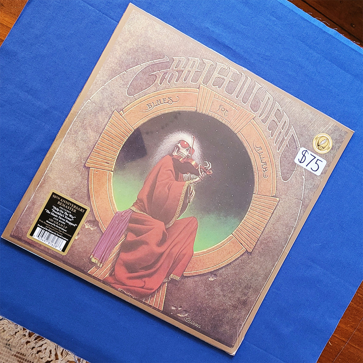 Grateful Dead 'Sanctuary' vinyl record album cover, photographed at an angle on a blue cloth surface. The cover art shows a robed figure seated in a circular frame, with the Grateful Dead logo at the top and a $75 price sticker in the upper right.