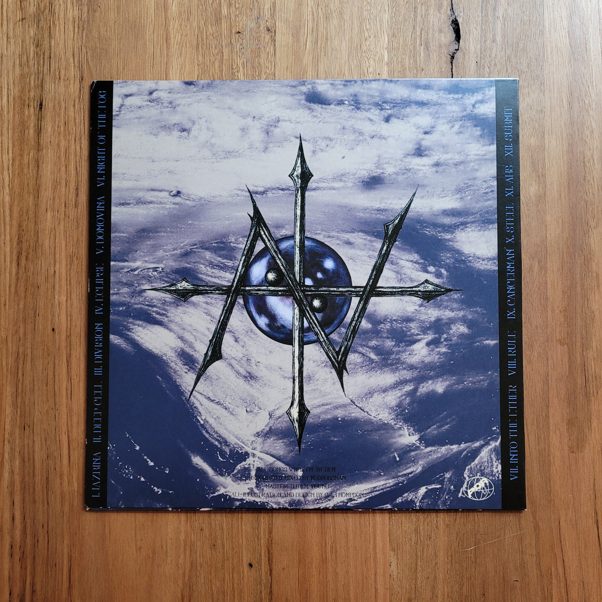 A square music album or book cover laid flat on a wooden surface, featuring a blue and white abstract landscape with a central black-and-silver geometric emblem and the letters 'AW' in the middle. The cover has black side bands with white text.