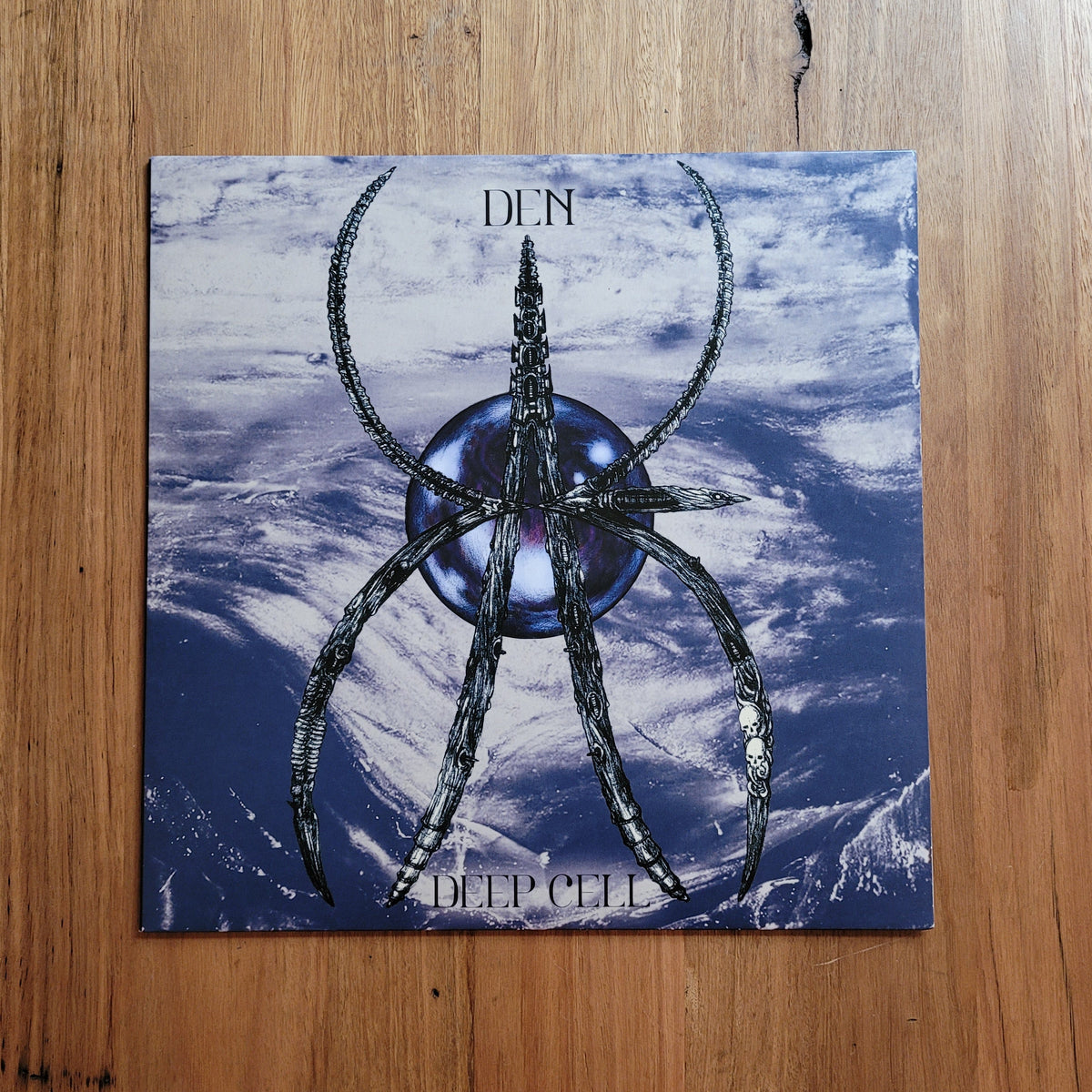 A square album or book cover titled 'DEEP CELL' with the word 'DEN' at the top, featuring a stylized metallic spider-like symbol over a blue and white cloud or icy background, placed on a wooden surface.