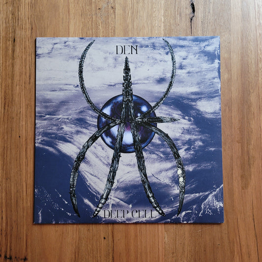 A square album or book cover titled 'DEEP CELL' with the word 'DEN' at the top, featuring a stylized metallic spider-like symbol over a blue and white cloud or icy background, placed on a wooden surface.