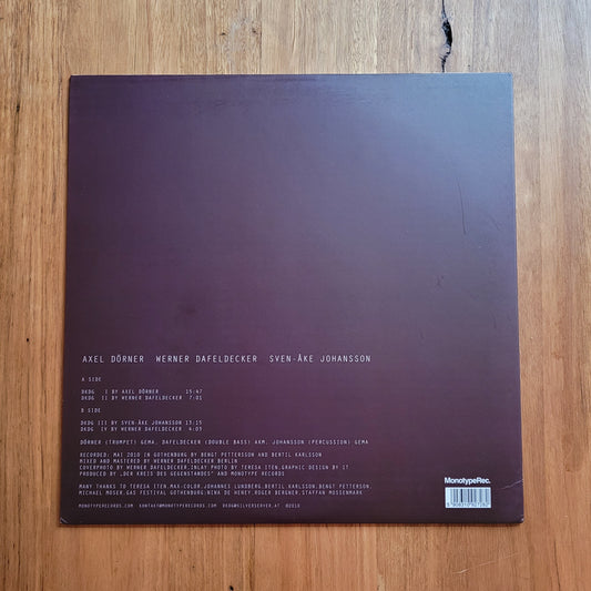 The back cover of a vinyl record album resting on a wooden surface. The cover is a very dark plum-purple color with white text listing the artist and track names, and a barcode in the bottom right corner.
