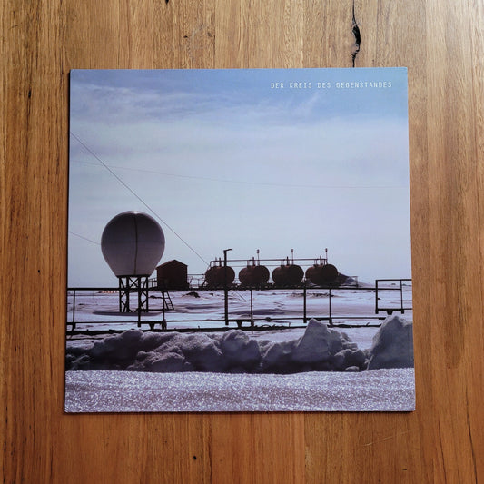 A square printed artwork or album cover placed on a wooden surface, showing a snowy industrial landscape with a large dome structure and several dark cylindrical tanks under a pale sky. Small text appears near the top right of the print.