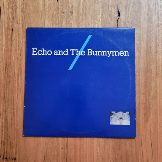 A square blue album cover for 'Echo and The Bunnymen' lies flat on a light brown wooden surface. The cover features white text and a light blue diagonal slash graphic, with a small square photo in the bottom right corner.