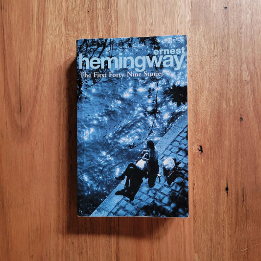 Ernest Hemingway - The First Forty-Nine Stories (Paperback) on a wooden surface with a serene backdrop.