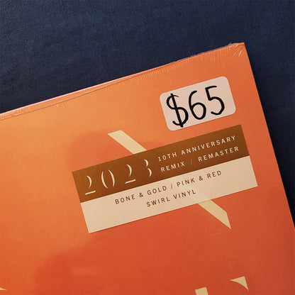 A close-up of a vinyl record sleeve for the 2023 10th Anniversary Remix / Remaster of Bone & Gold / Pink & Red / Swirl Vinyl. The sleeve is orange with brown and cream labels, wrapped in clear plastic, and a handwritten price sticker showing '$65' is placed on top.