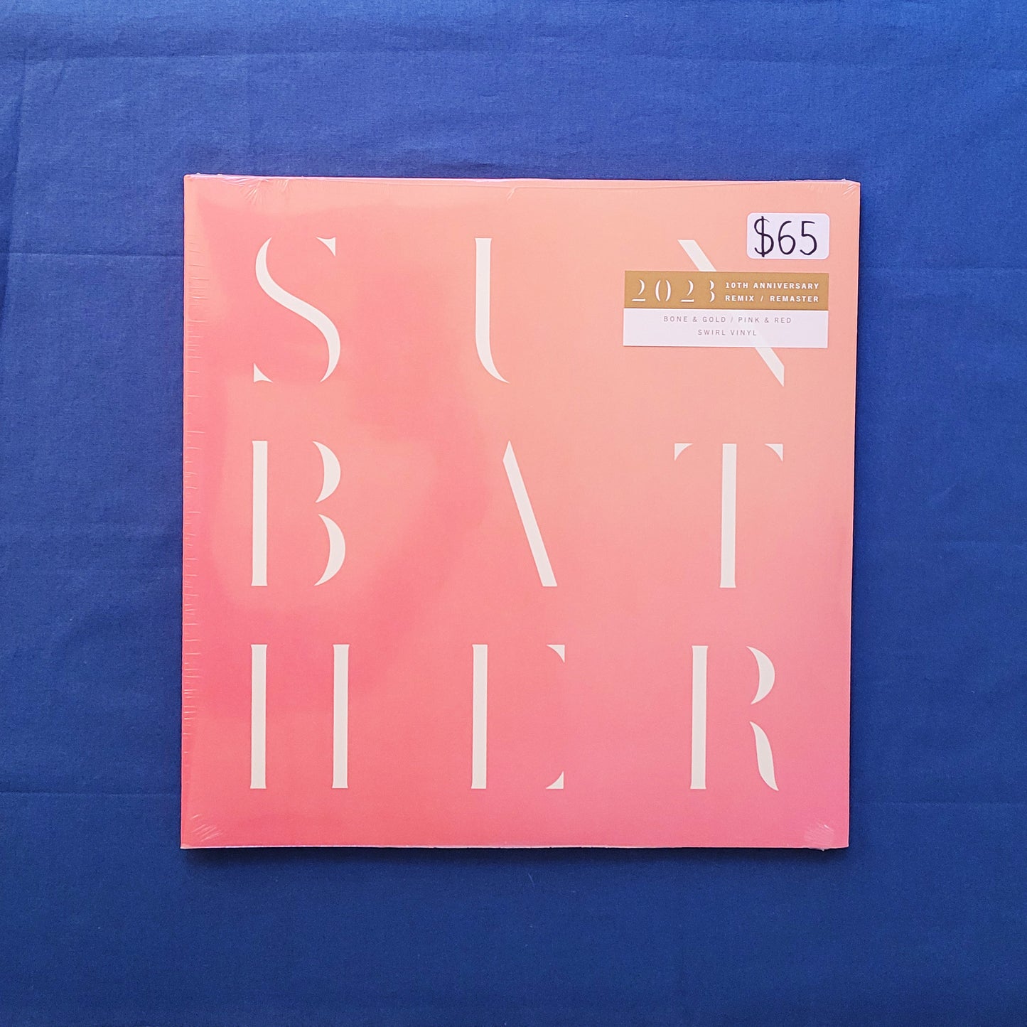 A square pink and peach album cover titled 'SUNBATHER' with large white typography, photographed flat against a blue fabric background. A small white price sticker reading '$65' is visible in the top right corner.