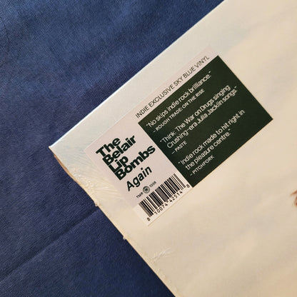 A close-up of a vinyl record sleeve or packaging corner on a blue textured fabric background. The white label reads 'The Belair Lip Bombs Again' with track listing text and a barcode, plus a dark green quote block.