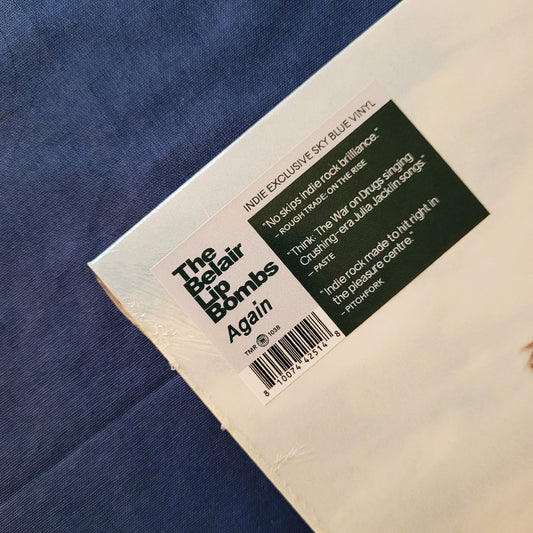A close-up of a vinyl record sleeve or packaging corner on a blue textured fabric background. The white label reads 'The Belair Lip Bombs Again' with track listing text and a barcode, plus a dark green quote block.