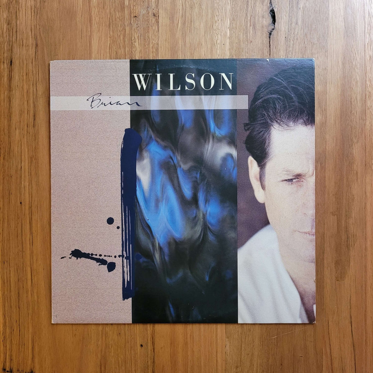 A square music album cover for Brian  Wilson's record lies flat on a wooden surface. The cover features a split design with a textured beige left side, a dark blue and black abstract brushstroke center, and a portrait of the artist on the right.