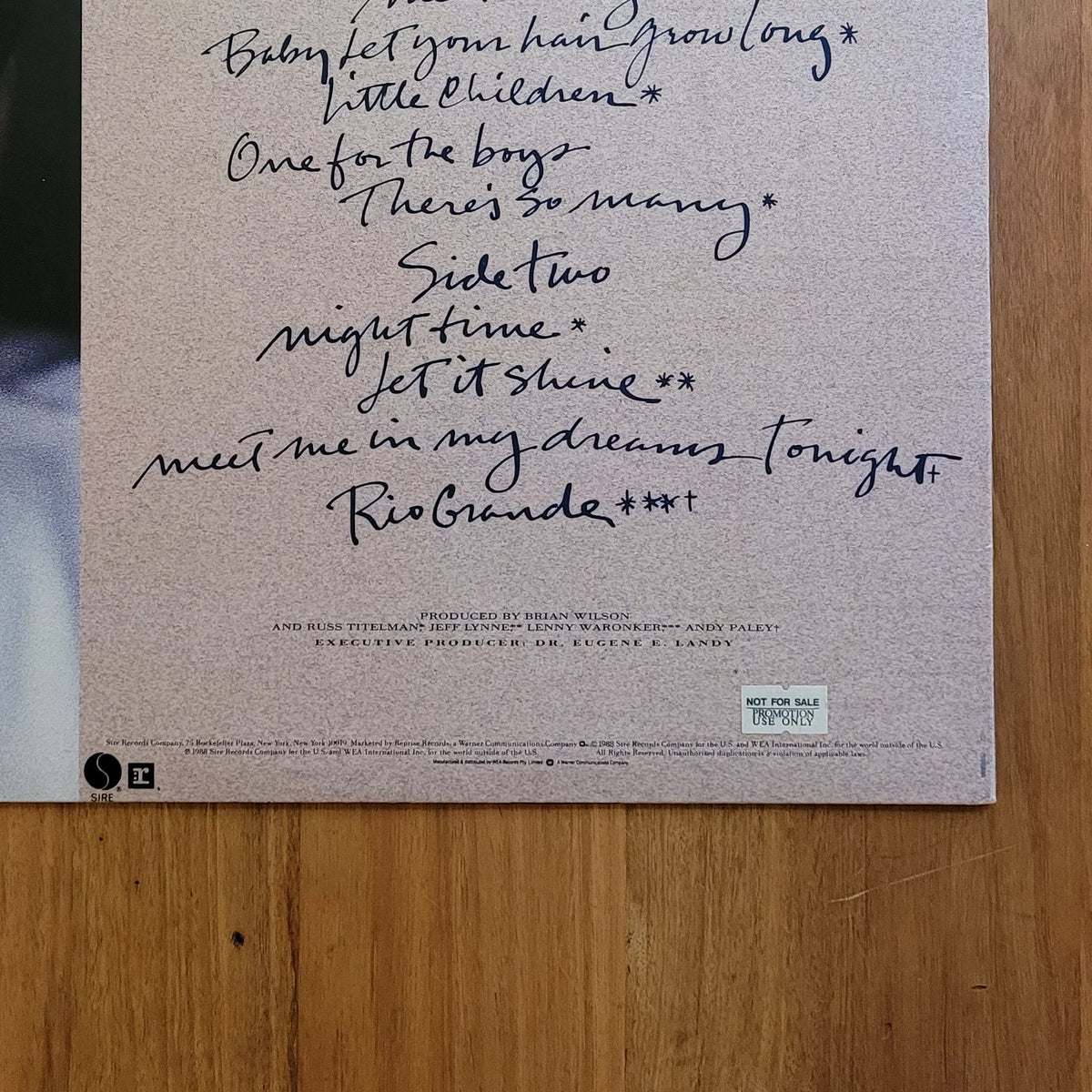 The back cover of a vinyl record album, featuring a handwritten tracklist and a partial portrait of a man on the left, displayed flat on a wooden surface.