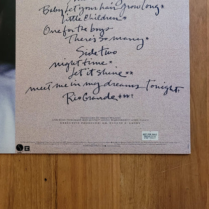The back cover of a vinyl record album, featuring a handwritten tracklist and a partial portrait of a man on the left, displayed flat on a wooden surface.