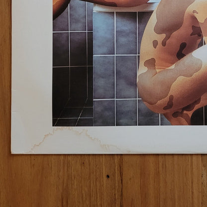 A square printed artwork or album cover titled 'FREEZE FRAME: GODLEY CREME' lies on a wooden floor. The cover features a stylized painting of two nude figures in front of blue tiled walls, with a wide white border around the artwork.