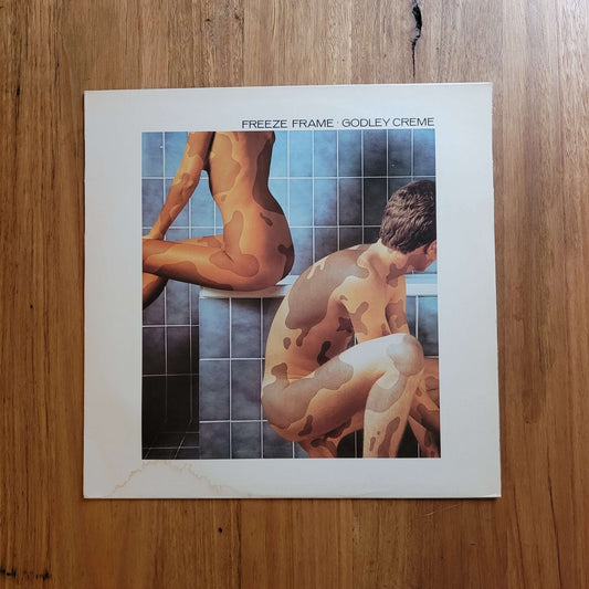 A square printed artwork or album cover titled 'FREEZE FRAME: GODLEY CREME' lies on a wooden floor. The cover features a stylized painting of two nude figures in front of blue tiled walls, with a wide white border around the artwork.