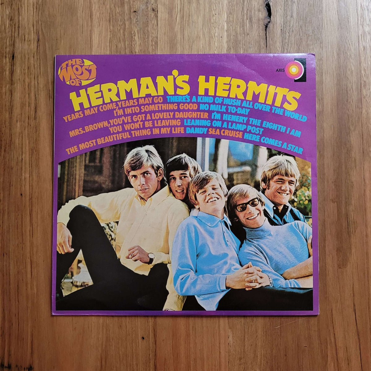 A vintage Herman's Hermits vinyl record album cover placed flat on a wooden surface. The cover features a purple border with yellow text and a photo of the five band members in colorful clothing.