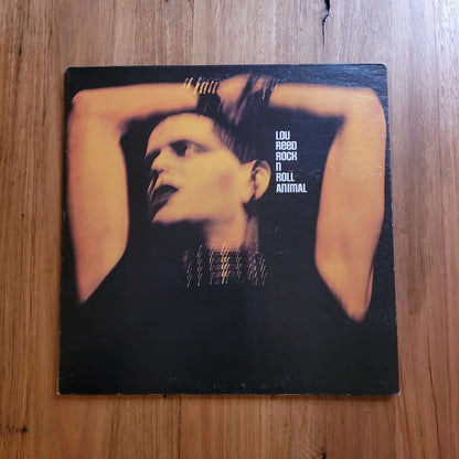 A vinyl record sleeve for Lou Reed's 'A Room with a View' resting on a wooden tabletop. The cover features a dramatic portrait of Lou Reed with his arms behind his head against a dark background, with the text 'LOU REED ROCK N' ROLL ANIMAL' printed on the right.